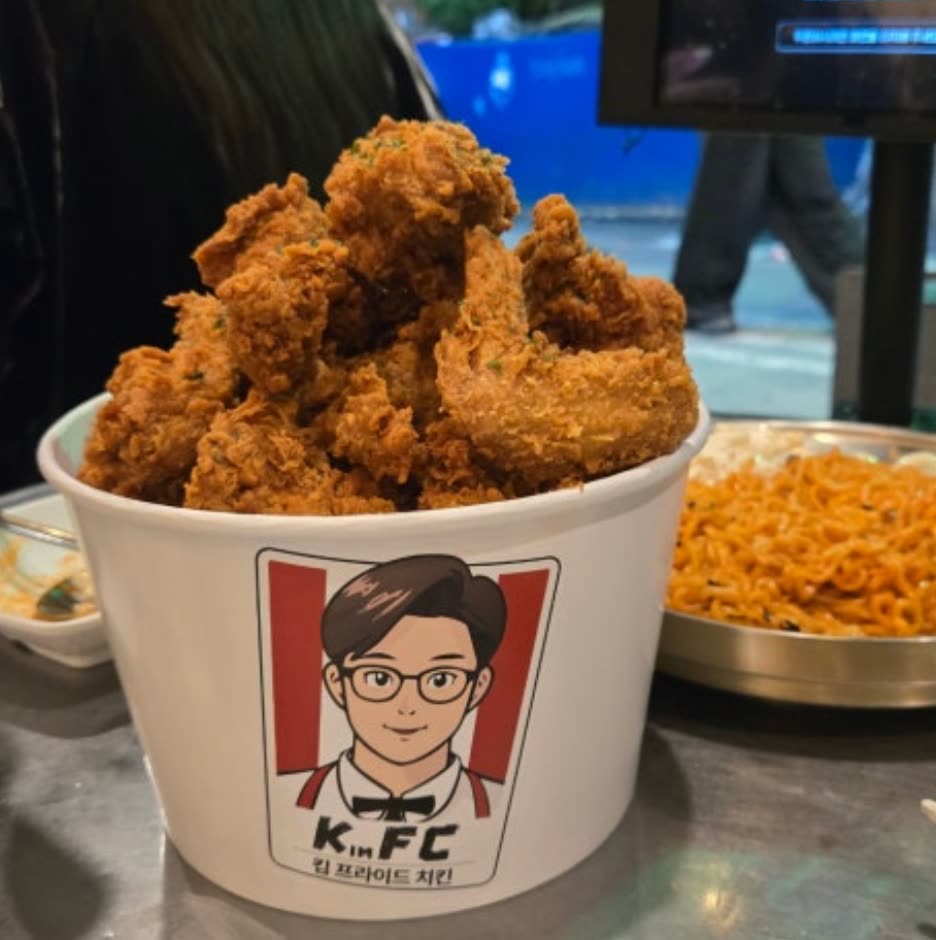 'KimFC 킴 프라이드 치킨' 로고가 붙은 흰 통에 수북이 담긴 뼈있는 후라이드치킨. 옆에는 볶음면이 함께 놓여 있다