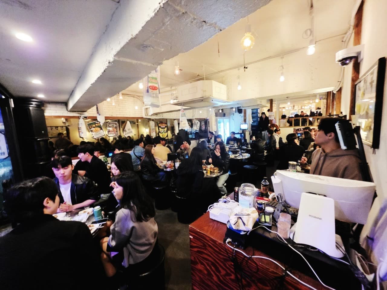 흰 벽과 노출 보 아래 둥근 테이블이 가득 찬 김실력포차 홀 내부. 거의 모든 자리에 손님이 앉아 있다